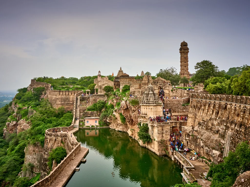 Vijay Stambh view at Chittorgarh Fort in Rajasthan under partly cloudy sky, featuring temples, crowds and reservoir, perfect Rajasthan tour package.