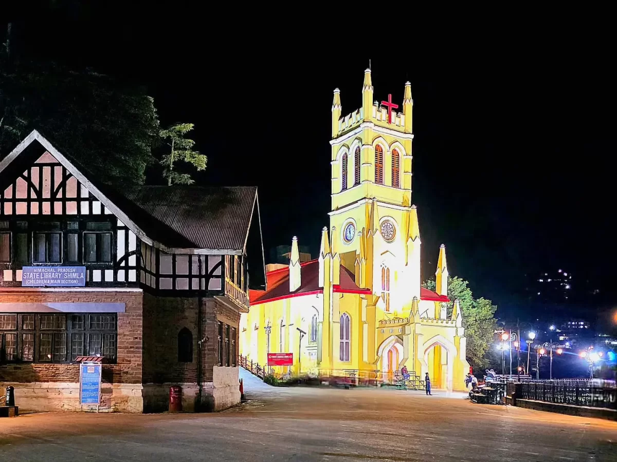 Christ Church Shimla Neo-Gothic yellow church tower illuminated at night with red cross adjacent colonial building trees city lights, perfect evening heritage view, Himachal Pradesh tour packages.