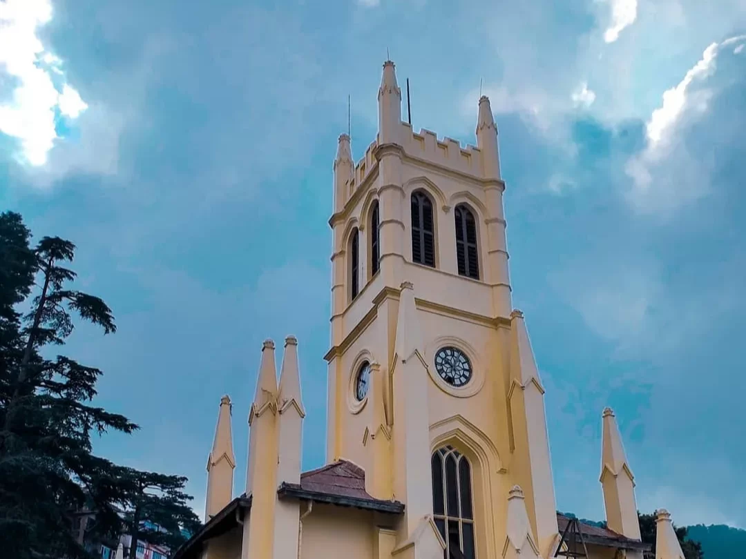 Christ Church Shimla iconic Neo-Gothic yellow clock tower with spires arched windows pine trees blue sky clouds, perfect colonial heritage landmark, Himachal Pradesh tour packages.