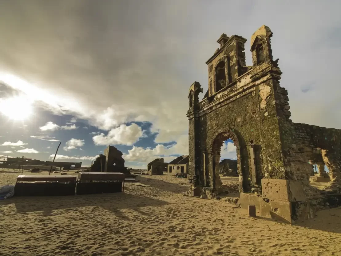 Church and Station Ruins in Dhanushkodi with sunbeams over crumbling stone arches and sandy remains under a cloudy sky.