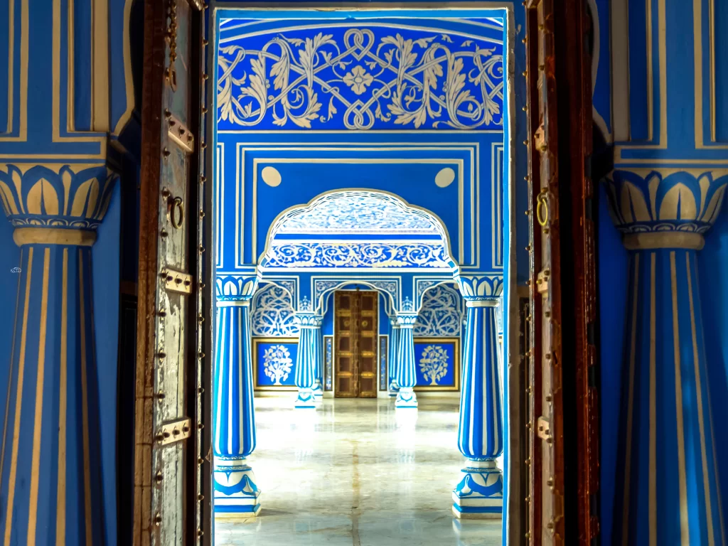 Iconic blue Rang Mandir corridor at City Palace Jaipur Rajasthan through massive wooden doors intricate white floral carvings pillars marble floor classic Rajasthani blue grandeur, perfect Rajasthan tour package.