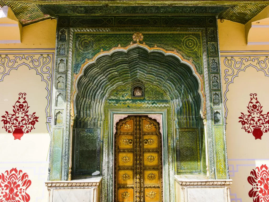 City Palace Jaipur Rajasthan elegant peacock green arched torana gateway with intricate floral tilework majestic carved wooden doors flanked by pink walls red floral motifs, opulent Rajasthani royal palace entrance heritage architectural tour package. 