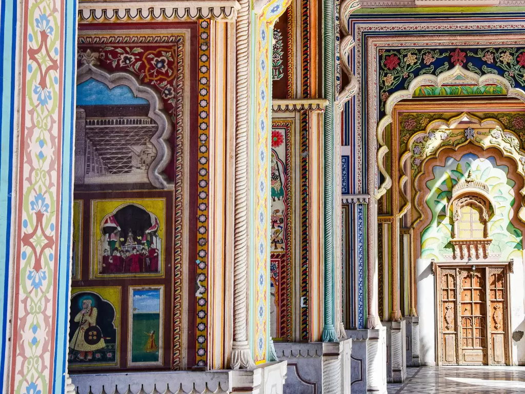 City Palace Jaipur Rajasthan vibrant corridor showcasing kaleidoscopic Rajasthani frescoes murals colorful arched jharokhas pillars intricate floral motifs wooden doors sunlight streaming, luxurious royal palace interior heritage architecture tour package
