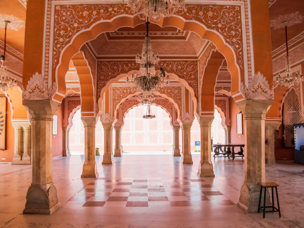 Opulent pink Diwan-i-Khas Mubarak Mahal hall City Palace Jaipur Rajasthan with multiple hanging chandeliers ornate arches checkered marble floor wooden table stools golden light, perfect Rajasthan tour package