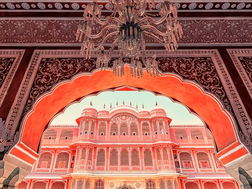 Grand Mubarak Mahal entrance archway framing Chandra Mahal tower City Palace Jaipur Rajasthan with multiple chandeliers intricate red carvings pink palace facade golden domes, perfect Rajasthan tour package.