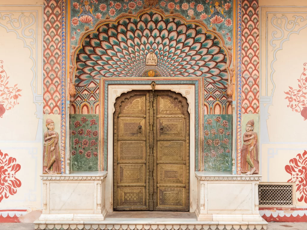 City Palace Jaipur Rajasthan stunning peacock turquoise fan-shaped jharokha archway with elaborate floral frescoes flanking gilded wooden doors pink walls guardian statues, exquisite Rajput palace gateway heritage architectural splendor tour package. 