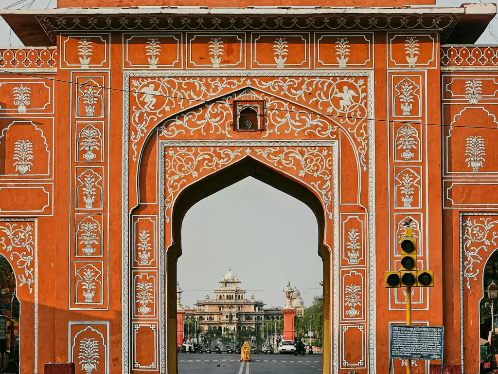 Majestic Tripolia Gate main entrance City Palace Jaipur Rajasthan with intricate orange terracotta carvings floral motifs framing Chandra Mahal domes in background traffic signal, perfect Rajasthan tour package