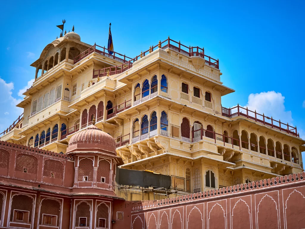 Majestic yellow Chandra Mahal tower at City Palace Jaipur Rajasthan multi-tiered balconies red lattice screens flags blue windows pink boundary walls under partly cloudy sky, perfect Rajasthan tour package