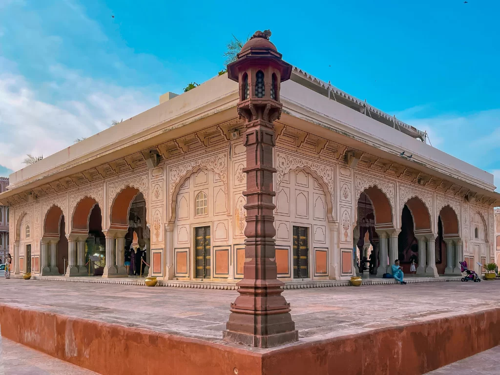 Classic Mubarak Mahal pavilion at City Palace Jaipur Rajasthan with red stone diya lamp intricate jharokhas arches tourists courtyard under clear blue partly cloudy sky, perfect Rajasthan tour package.