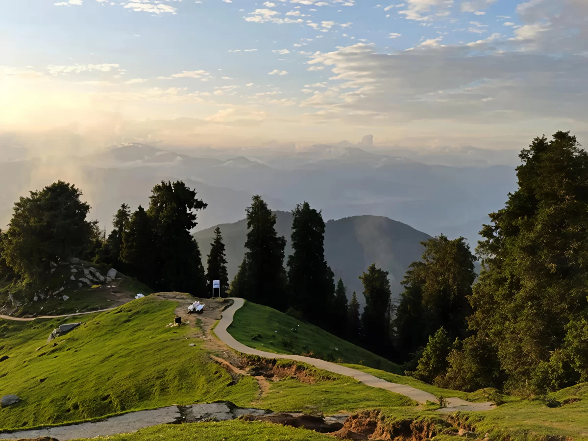 Trekking path to Dainkund Peak Dalhousie during sunset glow, featuring pine forests, misty Dhauladhar mountains, green meadows, perfect adventure Himachal Pradesh tour package.