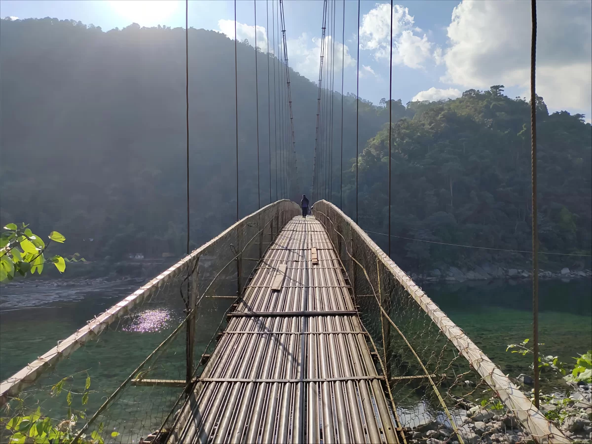 Dawki Suspension Bridge Dawki during sunny day, featuring bamboo hanging bridge person walking green hills turquoise Umngot River, perfect adventure experience Dawki Meghalaya tour package.