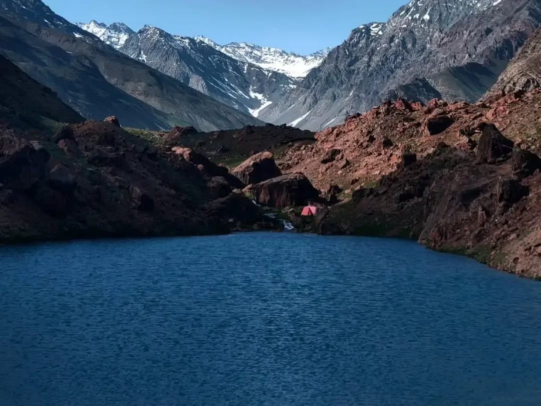 Deepak Tal Lahaul Spiti Himachal Pradesh India deep blue glacial lake framed towering snow capped peaks rugged red brown rocky mountains blue skies red tent lakeside camping foreground, perfect Manali Leh highway camping adventure tour package.