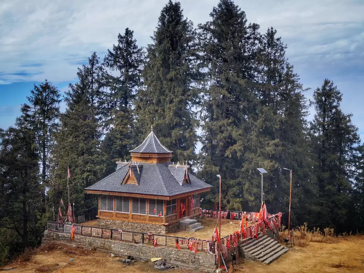Derthu Mata Temple in Narkanda featuring a traditional wooden Himachali temple with sloped roof, prayer flags along the boundary, stone platform, and tall deodar trees in the background.