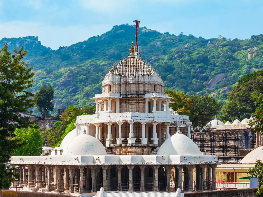 Dilwara Jain Temple Mount Abu, featuring intricate white marble multi-tiered shikhara domes pillars forested hills blue skies flag, perfect Rajasthan tour packages.