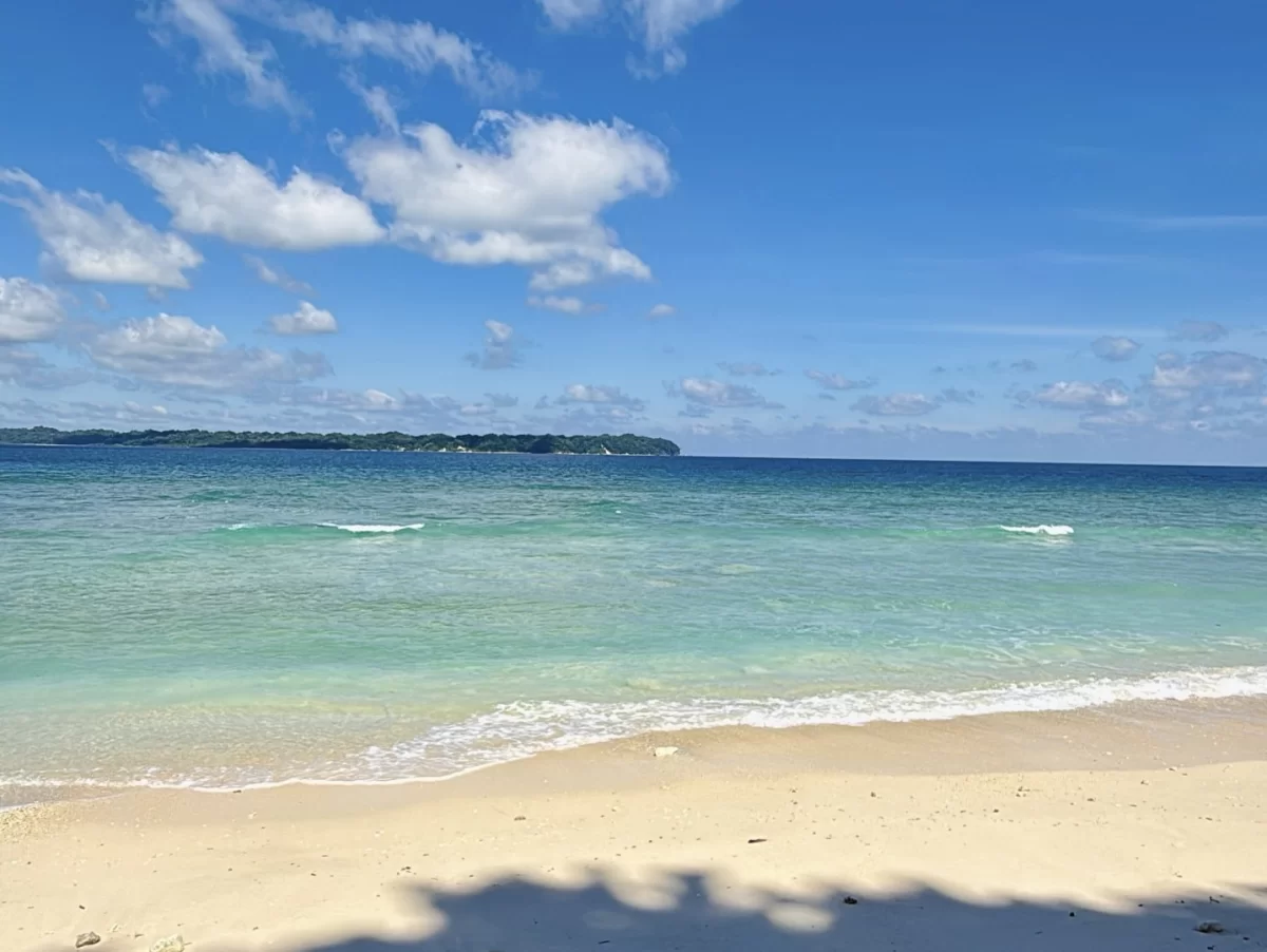 Panoramic Laxmanpur Beach Neil Island Andaman during sunny partly cloudy day, featuring white sands turquoise gradient lagoon offshore island palm shadows, perfect serene beach paradise Andaman tour package.