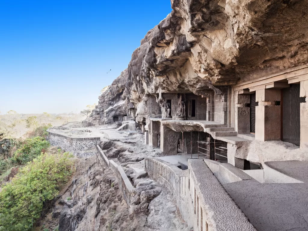 Rock-cut cave facades at Ellora Caves in Aurangabad during clear blue sky, featuring cliffside temples, greenery, perfect historical Aurangabad tour package. 