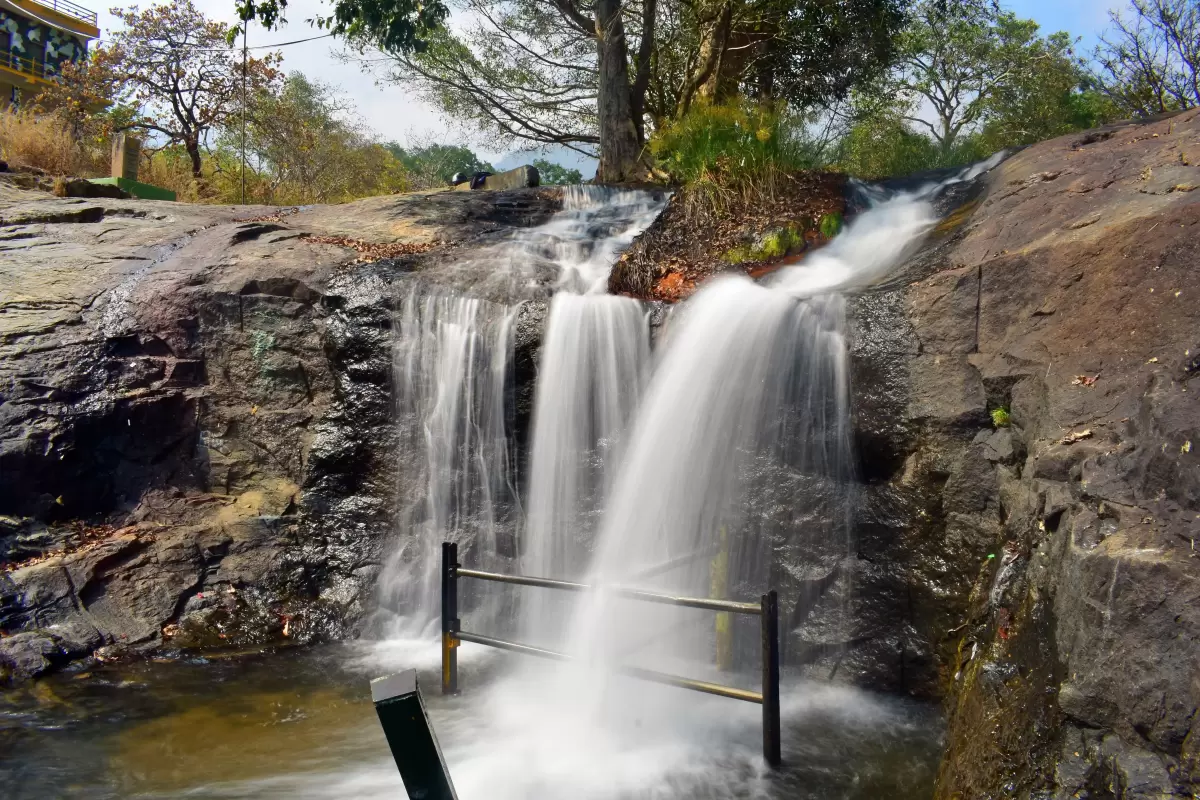 Kumbakkarai Falls at Kodaikanal foothills during sunny day, featuring cascading water, rocky pools, trees, railing, perfect adventure experience Tamil Nadu tour packages.