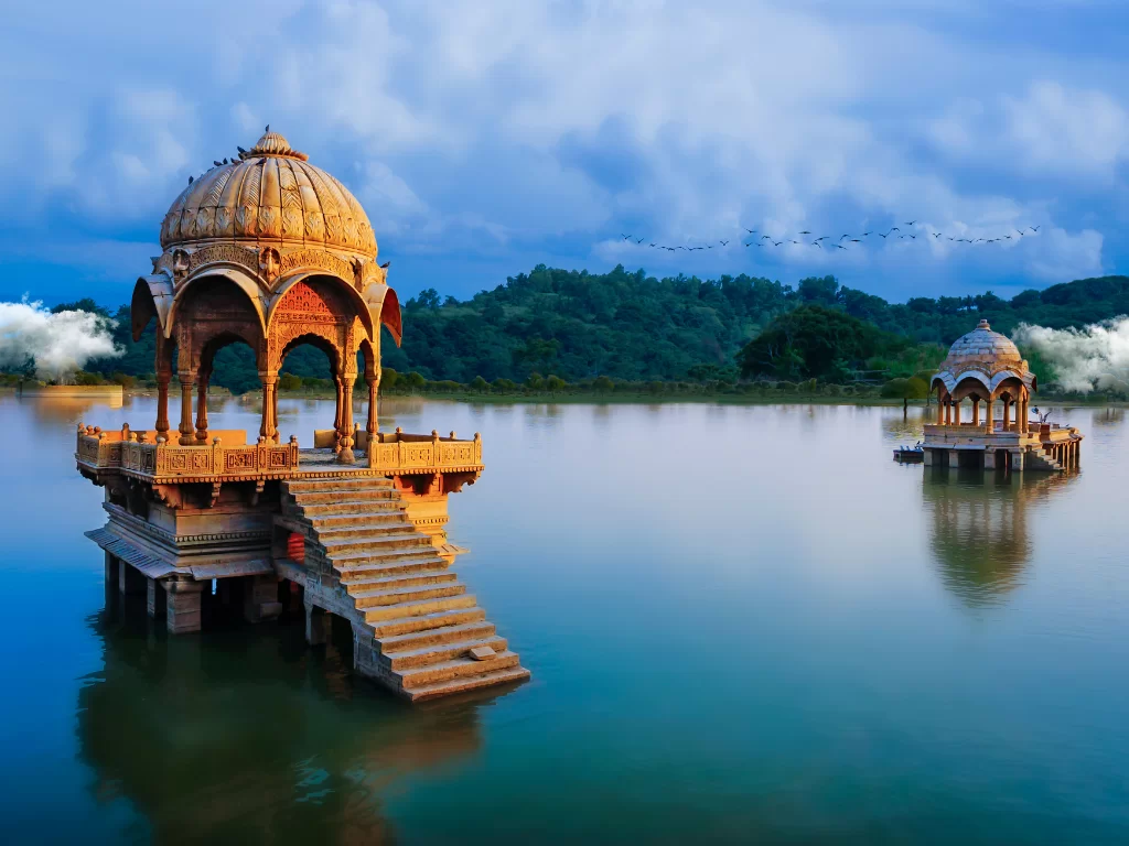 Gadisar Lake golden sandstone pavilion on lake pillars with stairs & birds under sunset clouds birds flying, perfect cultural heritage Jaisalmer Rajasthan tour packages.