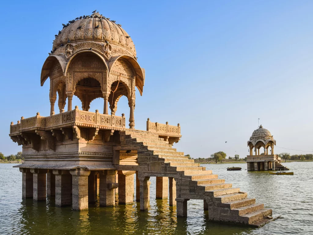 Gadisar Lake ornate sandstone pavilion on stilts with stairs amid waters & distant smaller pavilion under clear blue skies, perfect cultural heritage Jaisalmer Rajasthan tour packages. 