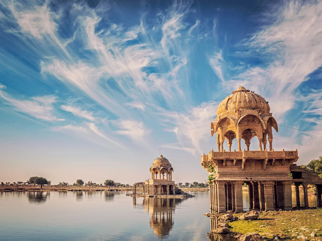 Gadisar Lake twin sandstone pavilions on lake islands under dramatic cloudy blue skies with reflections, perfect cultural heritage Jaisalmer Rajasthan tour packages.