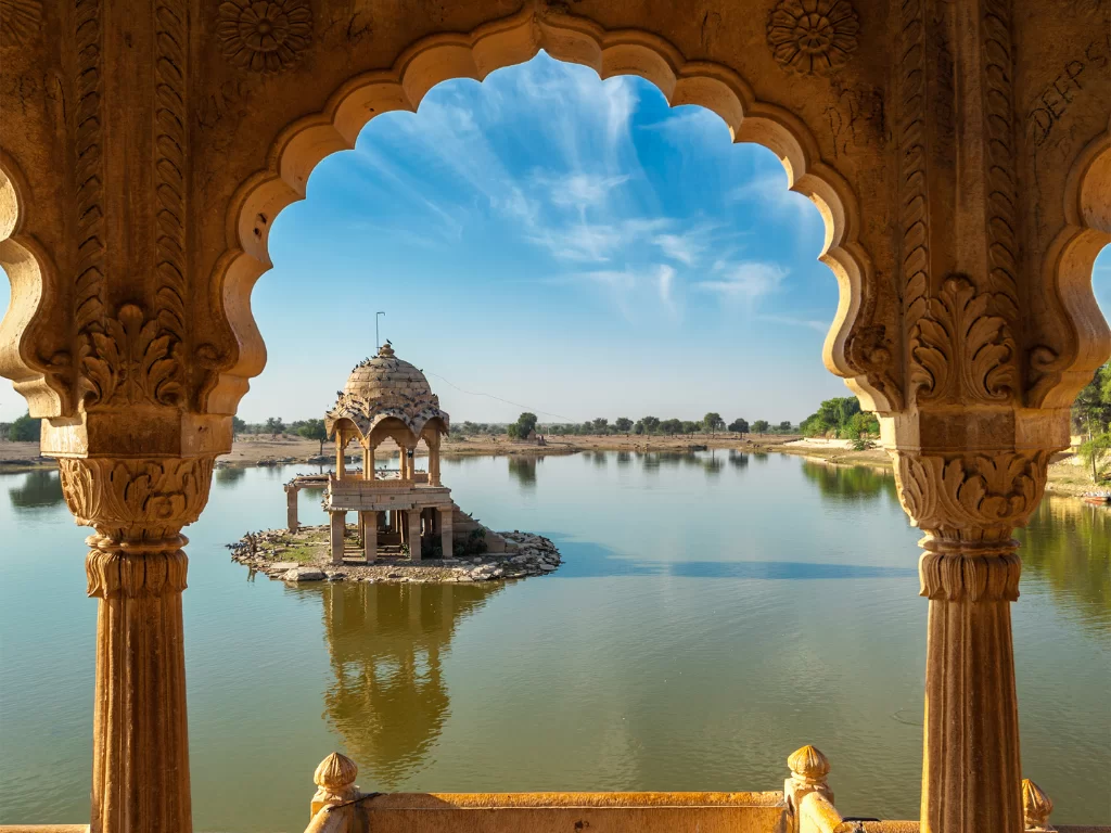 Gadisar Lake framed archway view of central sandstone pavilion on island amid serene waters blue skies, perfect cultural heritage Jaisalmer Rajasthan tour packages.