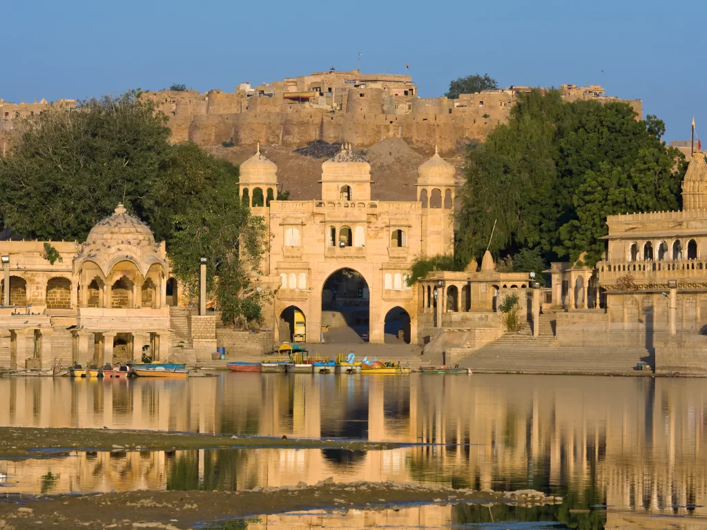 Gadisar Lake grand sandstone Tilon Gate entrance with pavilions, boats on reflecting water & Jaisalmer Fort backdrop, perfect cultural heritage Rajasthan tour packages.