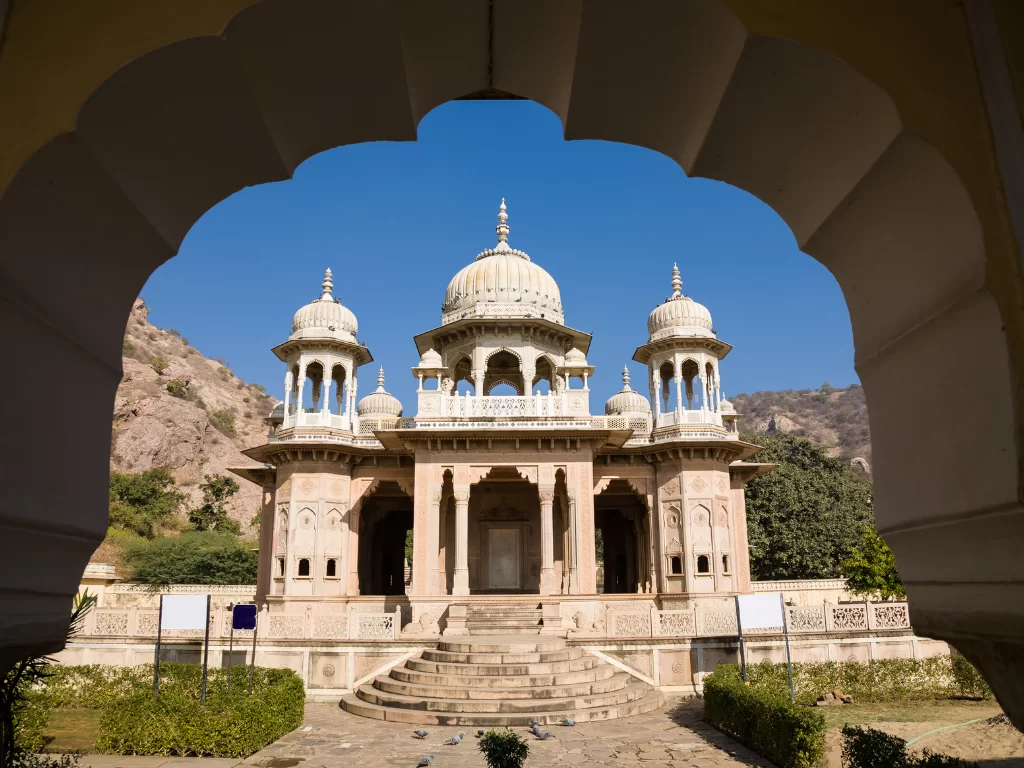 Gaitor Ki Chhatriyan Jaipur framed through stone archway pink sandstone multi-domed cenotaph pavilion intricate jali screens steps landscaped gardens Aravalli rocky hills backdrop blue sky trees, royal Rajput memorial architectural splendor heritage tour 