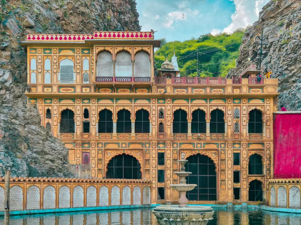 Galtaji Temple palace at Jaipur during partly cloudy day, featuring yellow architecture fountain hills greenery water pool, perfect spiritual Rajasthan tour package. 