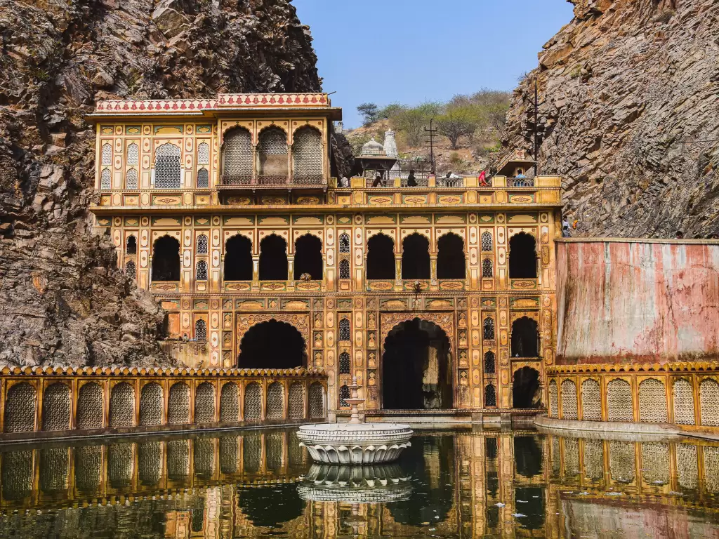 Galtaji Temple at Jaipur hills during clear skies, featuring yellow palace arches reflection pool rocks greenery, perfect spiritual Rajasthan tour package. 