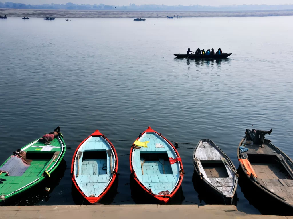 Colorful boats docked at Ganges River Varanasi during morning mist, featuring distant ghats rowers, perfect adventure Uttar Pradesh tour package.