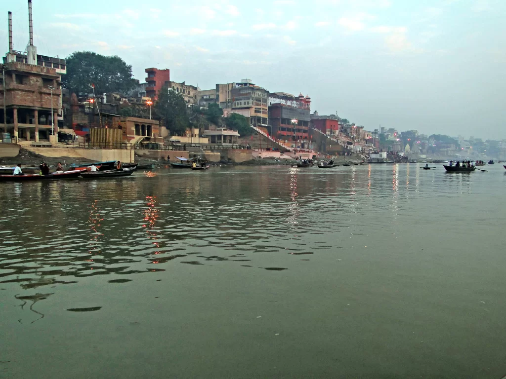 Ganges River Varanasi evening, boats ghats buildings trees lights misty water reflections colorful skyline, perfect Uttar Pradesh tour package.