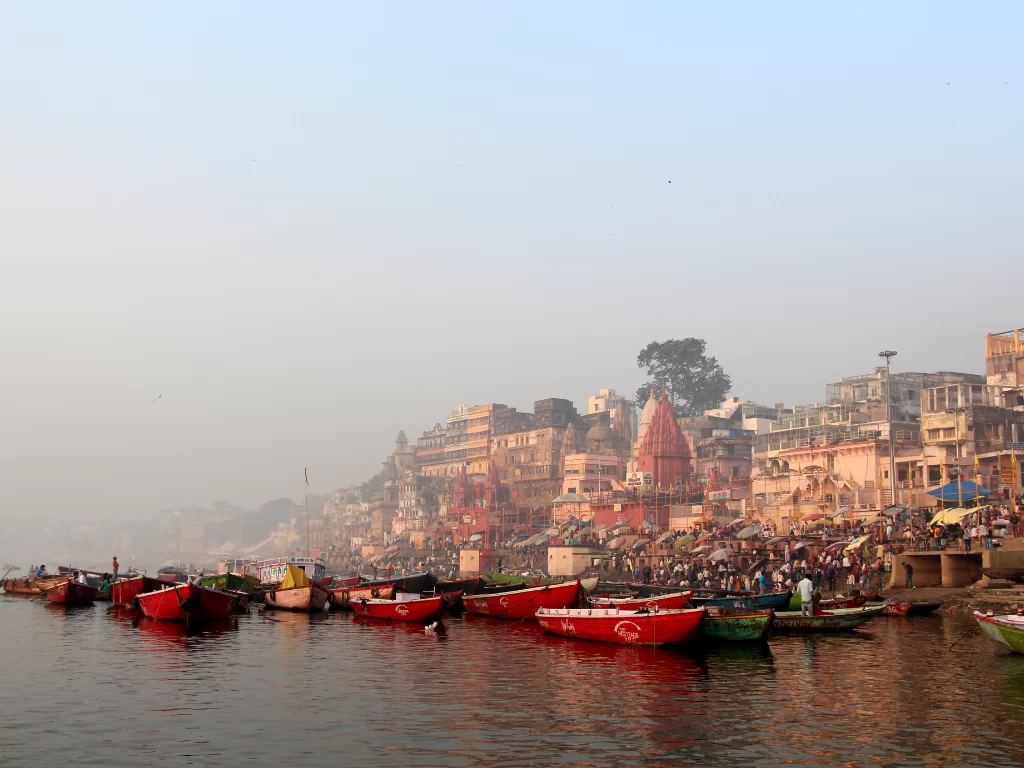 Red boats at Ganges River Varanasi during misty morning, featuring temples crowds reflections, perfect adventure Uttar Pradesh tour package.