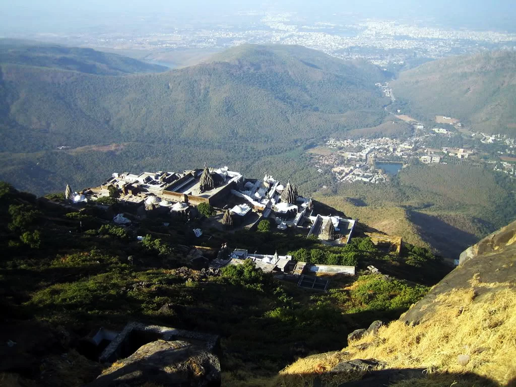 Girnar Jain temples at Junagadh during clear day, featuring temple cluster on hilltop amid mountains, city views, perfect pilgrimage experience with Gujarat tour packages.