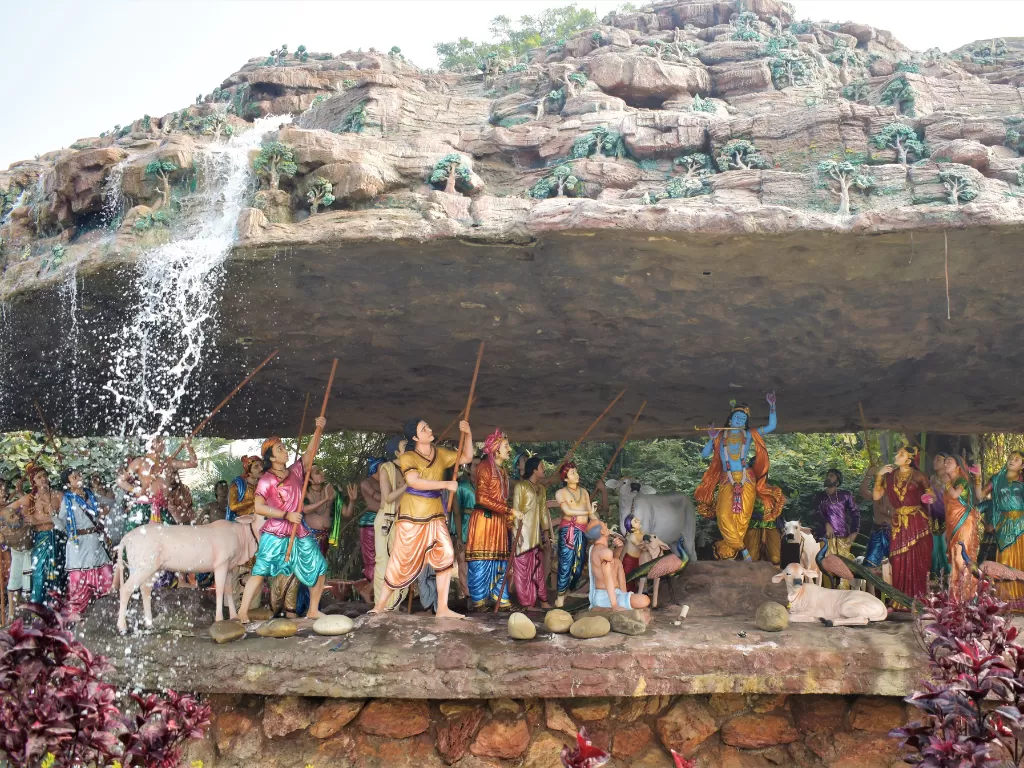 Krishna lifting Govardhan Hill at Govardhan Hill Mathura during clear daytime, featuring colorful statues, cascading waterfall, cows, perfect spiritual experience with Mathura Vrindavan tour package