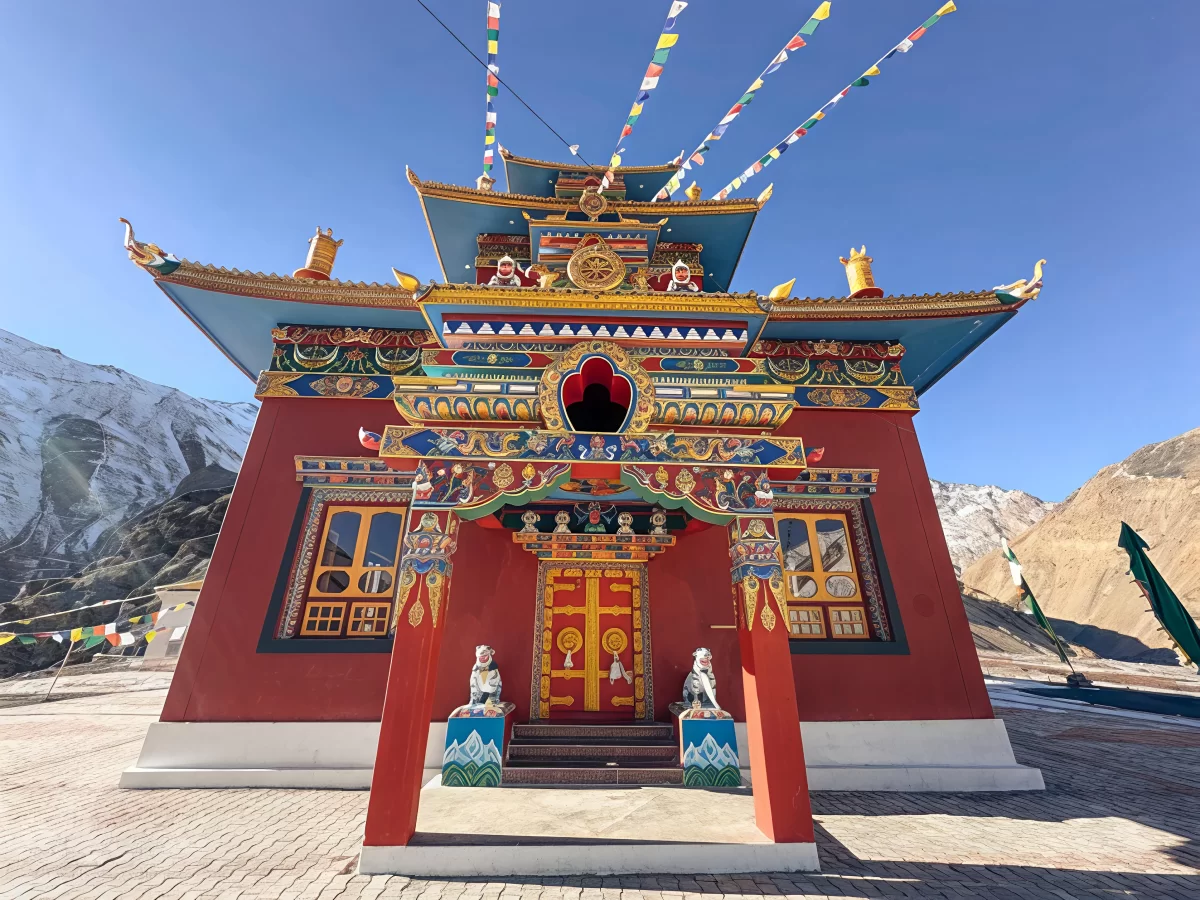 Gue Monastery Kaza Spiti close-up winter, featuring vibrantly painted maroon temple golden roofs prayer flags snowy terrain distant mountains blue skies, perfect Himachal Pradesh tour packages.