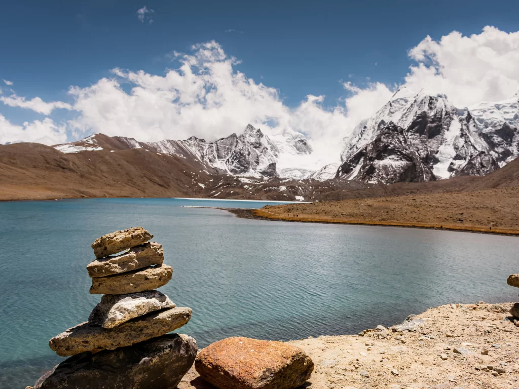 Prayer flags and stone cairns at Gurudongmar Lake North Sikkim during clear skies, featuring turquoise waters snow mountains, perfect spiritual adventure North Sikkim tour package.
