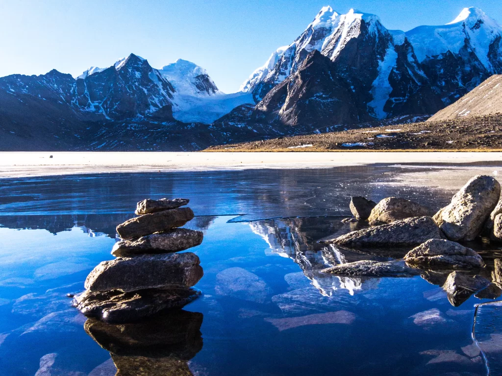 Gurudongmar Lake during clear daylight, featuring stone cairns reflecting on icy surface with snow peaks, perfect nature Sikkim tour package.