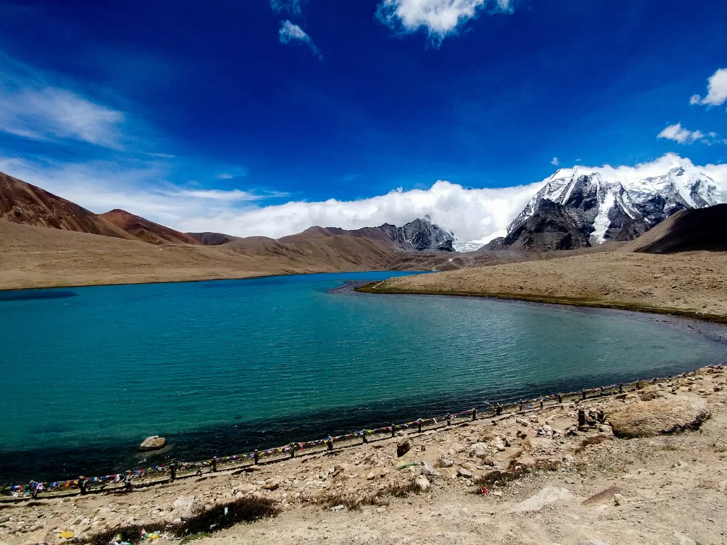 Gurudongmar Lake at North Sikkim during clear skies, featuring turquoise waters snow-capped peaks prayer flags, perfect spiritual adventure North Sikkim tour package.