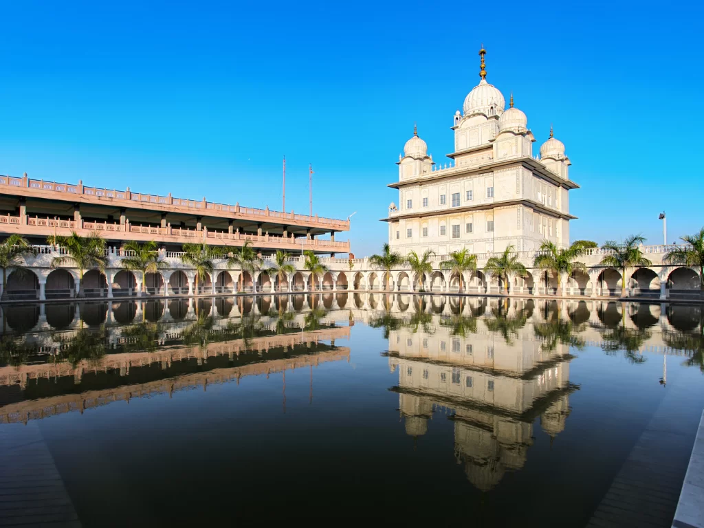 Gurdwara Data Bandi Chhor Sahib Gwalior during clear afternoon, featuring white marble domes reflected in sarovar with arched colonnades and palm trees, perfect heritage experience in Madhya Pradesh tour package.