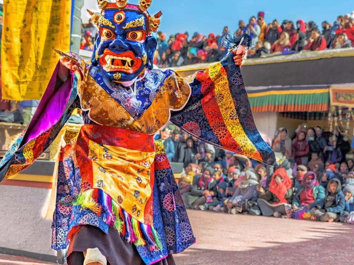 Blue-faced demon Cham dancer at Gustor Festival Spituk Monastery Ladakh during clear skies, featuring multicolored silk robes prayer flags crowd, perfect cultural adventure experience with Ladakh tour package.