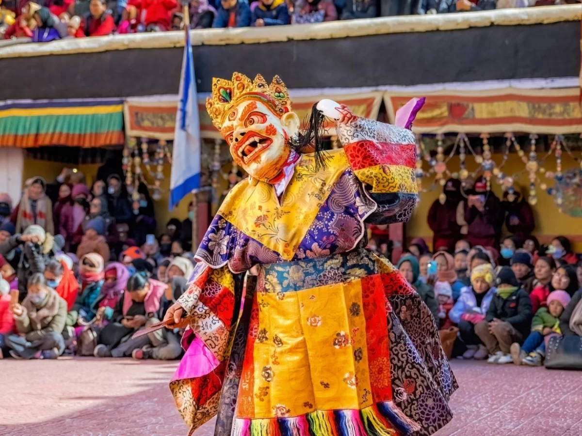 Golden-faced Cham dancer performing at Gustor Festival Spituk Monastery Ladakh during sunny day, featuring multicolored robes crowd temple backdrop, perfect cultural adventure experience with Ladakh tour package