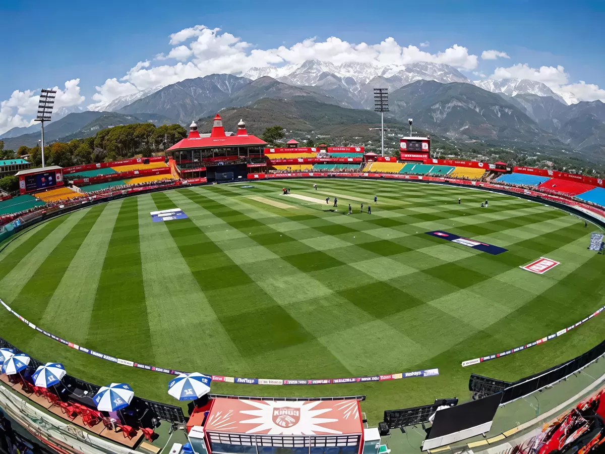 HPCA Stadium Dharamshala featuring lush green cricket ground, colorful stands, and snow-capped Dhauladhar mountains in the backdrop.