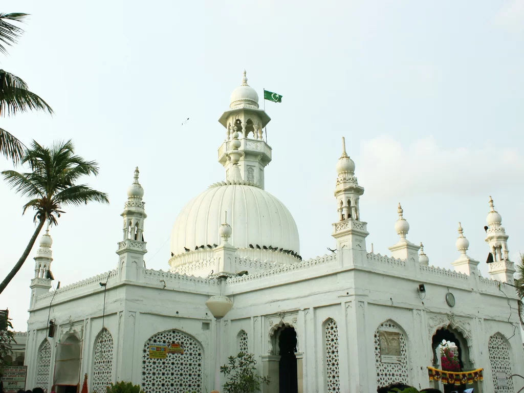 Haji Ali Dargah Mumbai full facade with white dome minarets and Pakistan flag amid palm trees, perfect Mumbai spiritual heritage tour package. 