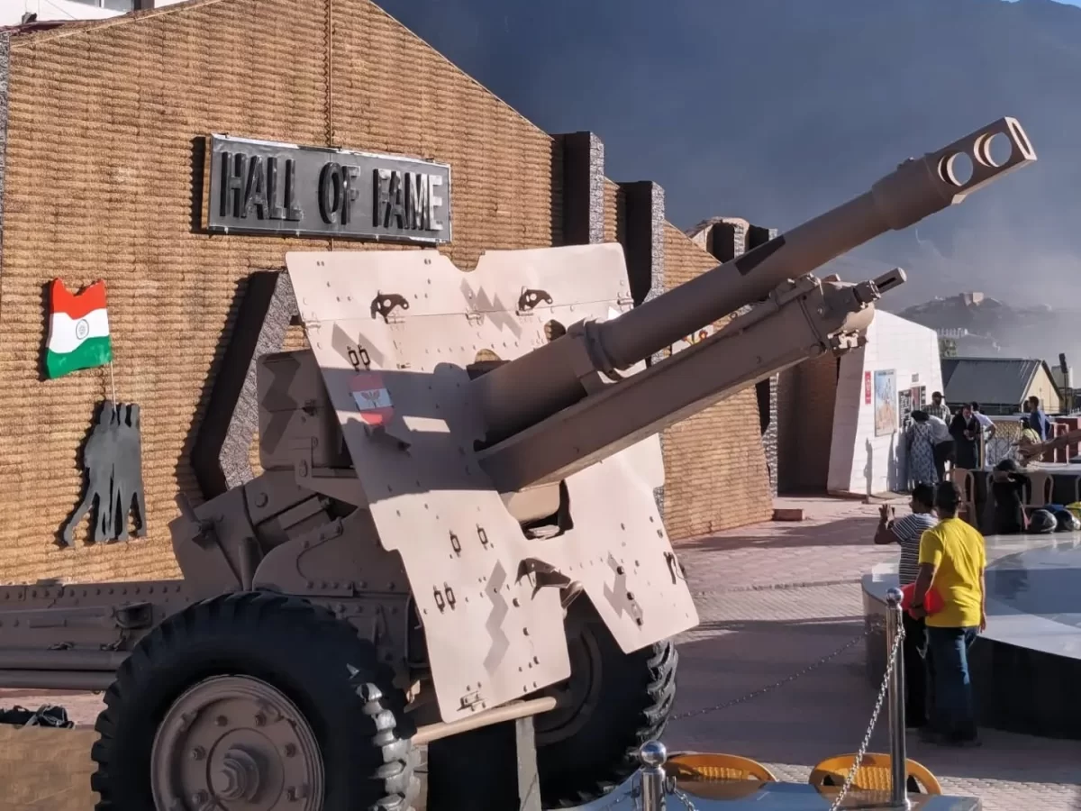 Hall of Fame Leh Ladakh captured Pakistani howitzer artillery gun tan camouflage wheels barrel signboard brick wall tourists yellow shirt cloudy sky military trophy display.