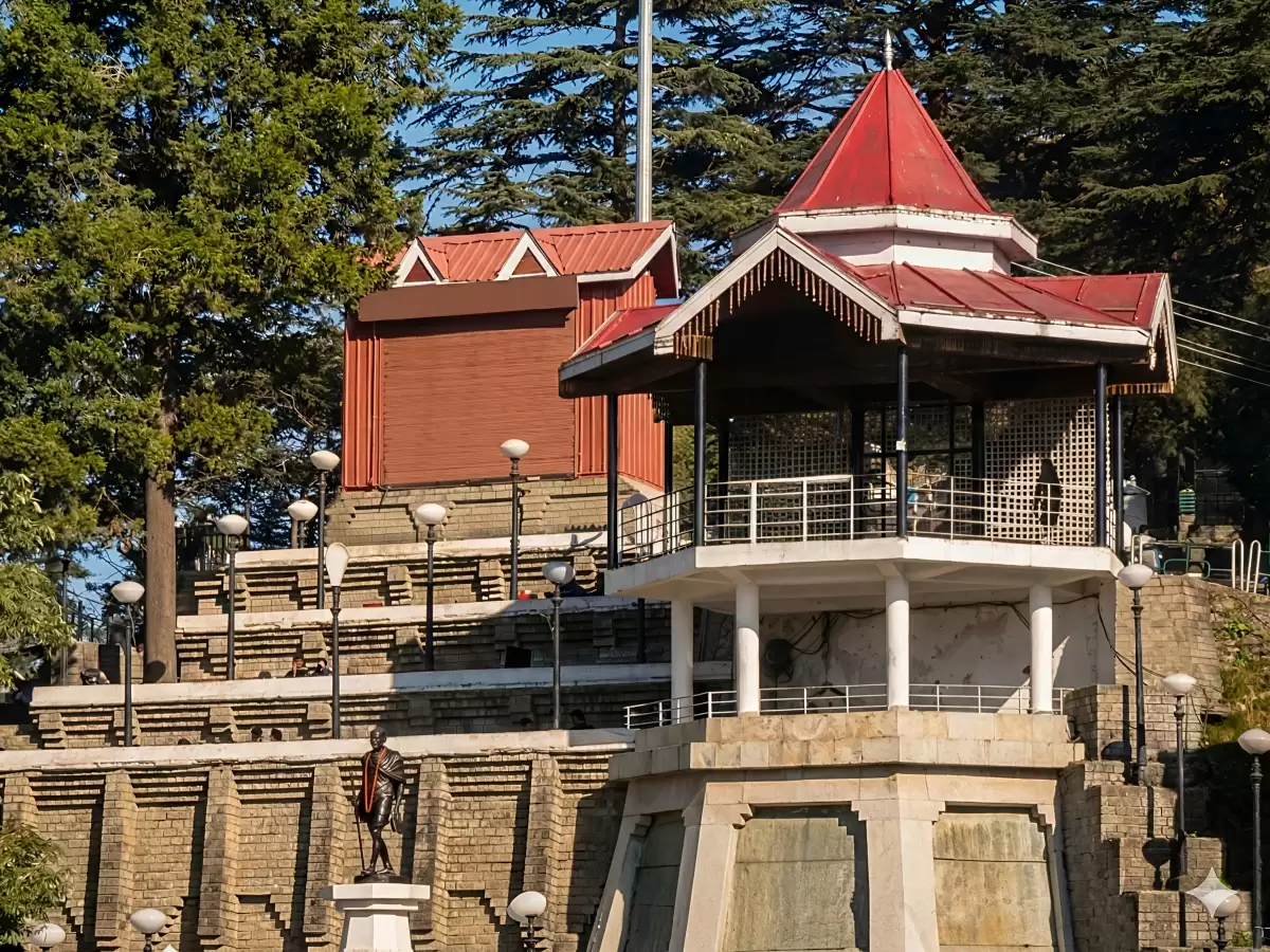 Hawa Ghar in Shangarh featuring a pavilion-style structure with a red sloping roof, stone terraces, surrounding deodar trees, and a statue in the foreground set against a hillside backdrop.
