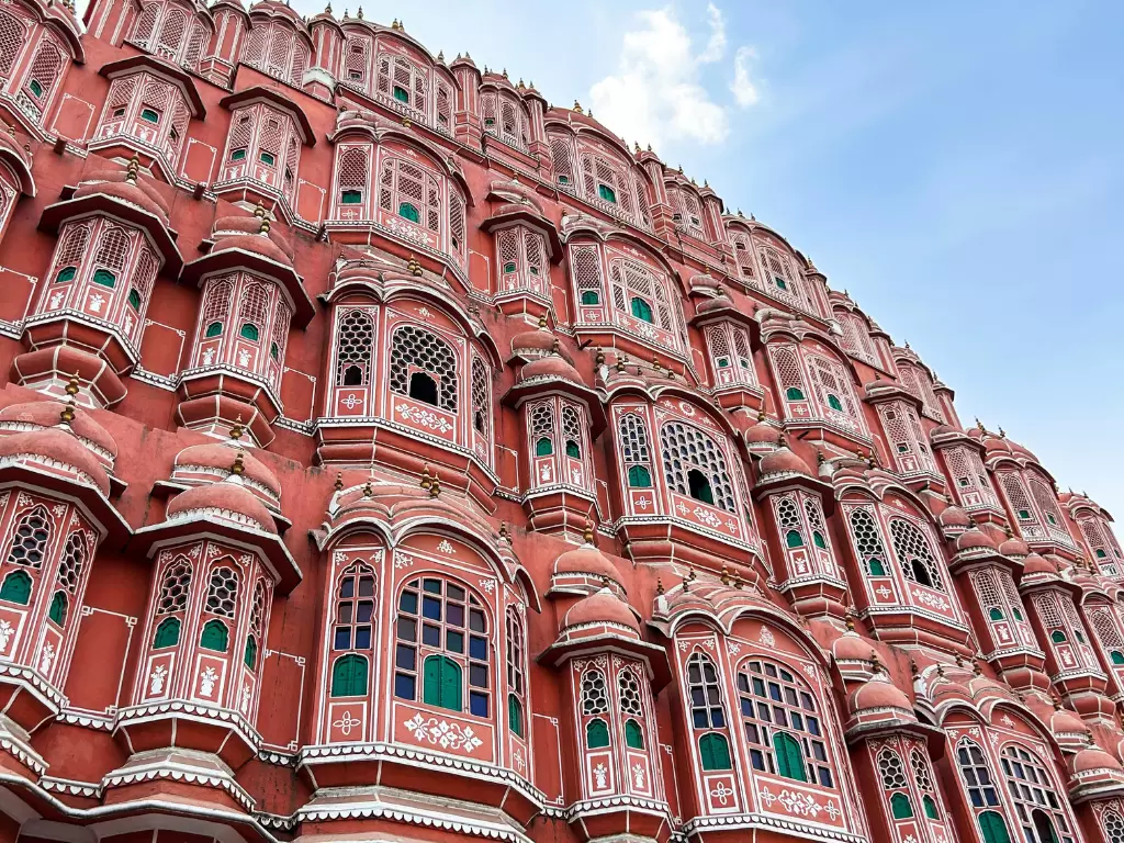 Hawa Mahal low angle Jaipur partly cloudy, featuring pink jharokhas green windows domes blue sky, perfect romantic experience Rajasthan tour packages.