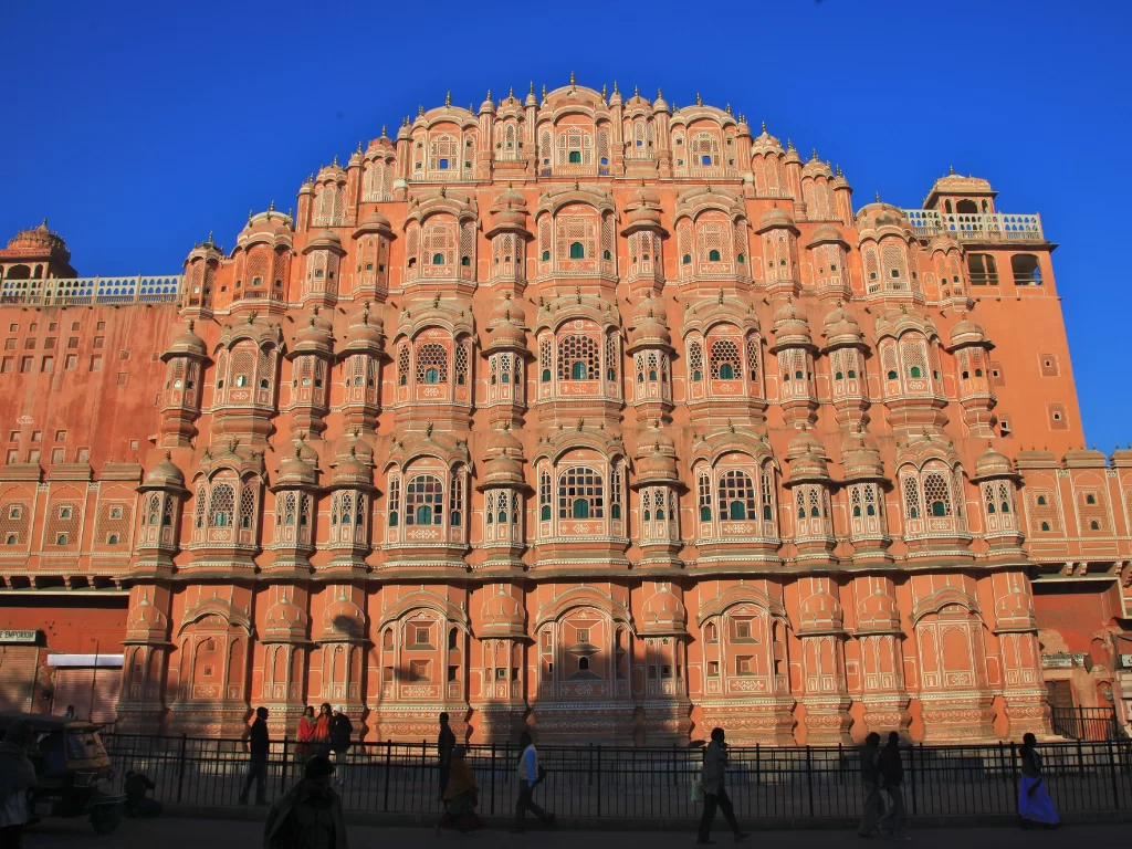 Hawa Mahal Jaipur sunny day, featuring pink facade tourists shadows street blue sky, perfect romantic experience Rajasthan tour packages.