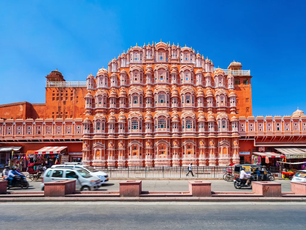 Hawa Mahal Jaipur sunny day, featuring pink palace cars rickshaws street crowds blue sky, perfect romantic experience Rajasthan tour packages.