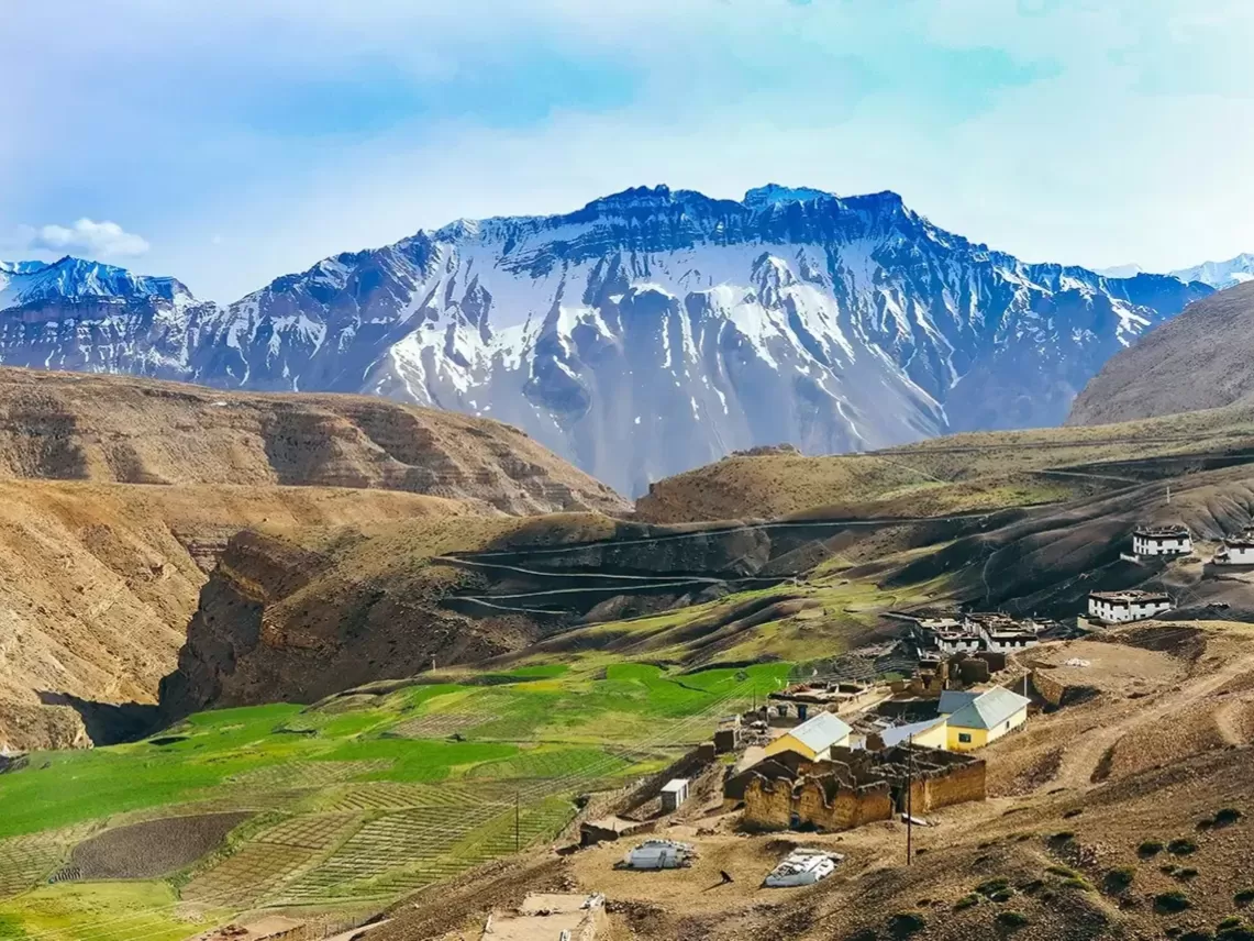 Hikkim Village Spiti near Kaza during clear skies, featuring traditional houses green fields rocky valley snow mountains, perfect cultural experience Himachal tour package.