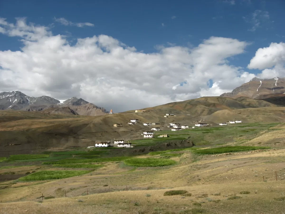 Hikkim Village Spiti near Kaza during partly cloudy skies, featuring white houses green fields brown hills snow mountains, perfect cultural experience Himachal tour package.
